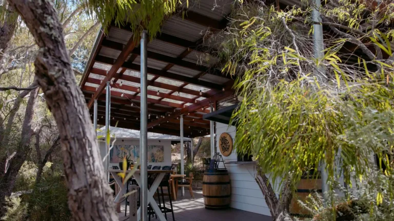 Exterior view of Valle du Venom cellar door nestled among native bushland in Yallingup, featuring a timber and steel pergola with festoon lights and wine barrel decor