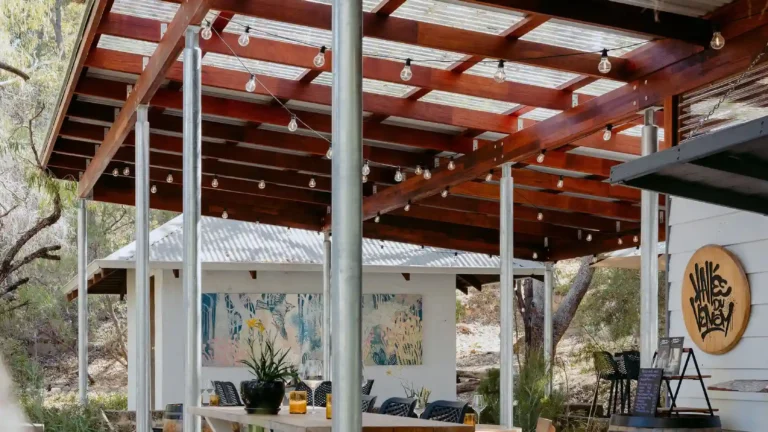 Covered outdoor deck at Valle du Venom winery in Yallingup with timber pergola, festoon lighting, long bar-height dining table, and wine barrels alongside native bushland
