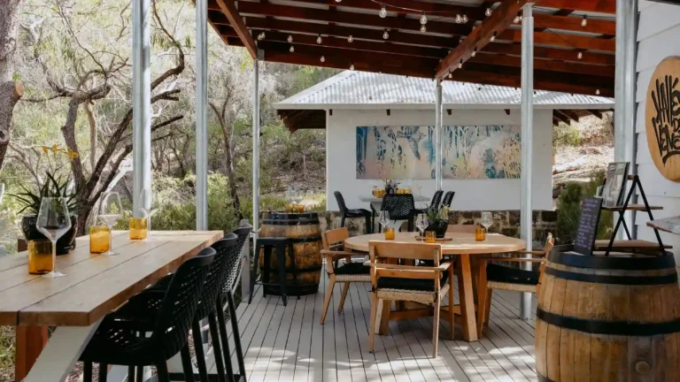 Full view of Valle du Venom winery deck in Yallingup with bar-height seating, round dining tables, wine barrels, festoon lights, and colourful artwork displayed on the back wall